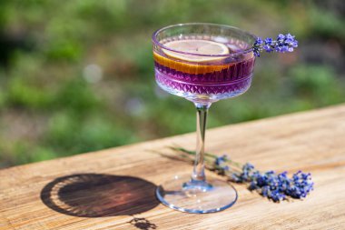 Yeşil bir yaz bahçesine karşı ahşap masada zarif kristal bardakta servis edilen parlak kokteyl ya da limonata. Eflatun çiçekli mor renkli içecek ve bir dilim limon.