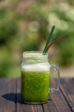 Cam bardakta taze yeşil meyve püresi. Yazlık bahçe arka planında metal kamışla ahşap masada, yakın plan. Sağlıklı beslenme kavramı. Yapraklı yeşillerden, otlardan, sebzelerden ve meyvelerden arınma içeceği