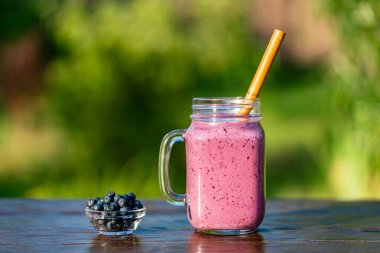 Cam bardakta taze yabanmersini püresi ve bambu kamışla ahşap masada, yaz bahçesi arka planında, yakın çekim. Sağlıklı beslenme kavramı. Yabanmersini, muz, bal ve yoğurttan meyve suyu.