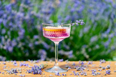 Yeşil bir yaz bahçesine karşı ahşap masada zarif kristal bardakta servis edilen parlak kokteyl ya da limonata. Eflatun çiçekli mor renkli içecek ve bir dilim limon.