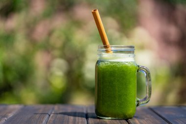Cam bardakta taze yeşil smoothie bambu kamışla yaz bahçesi arka planına karşı ahşap masada, yakın çekim. Sağlıklı beslenme kavramı. Yapraklı yeşillerden, otlardan, sebzelerden ve meyvelerden arınma içeceği