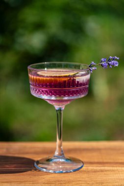 Yeşil bir yaz bahçesine karşı ahşap masada zarif kristal bardakta servis edilen parlak kokteyl ya da limonata. Eflatun çiçekli mor renkli içecek ve bir dilim limon.