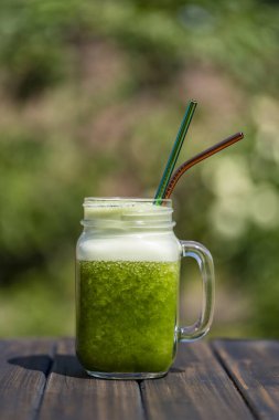 Cam bardakta taze yeşil meyve püresi. Yazlık bahçe arka planında metal kamışla ahşap masada, yakın plan. Sağlıklı beslenme kavramı. Yapraklı yeşillerden, otlardan, sebzelerden ve meyvelerden arınma içeceği