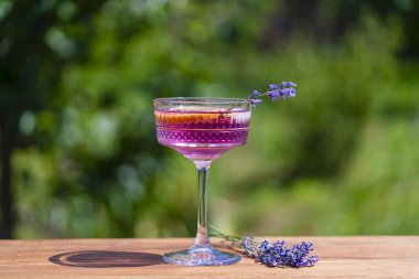 Yeşil bir yaz bahçesine karşı ahşap masada zarif kristal bardakta servis edilen parlak kokteyl ya da limonata. Eflatun çiçekli mor renkli içecek ve bir dilim limon.