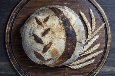Ahşap kesim tahtası arka planında buğday kulaklı taze ekşi ekmeğin yuvarlak somunu, yakın plan, üst manzara. Rustik ekşi ekmek. Evde sağlıklı ekşi ekmek pişirmek.