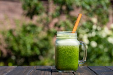 Cam bardakta taze yeşil smoothie bambu kamışla yaz bahçesi arka planına karşı ahşap masada, yakın çekim. Sağlıklı beslenme kavramı. Yapraklı yeşillerden, otlardan, sebzelerden ve meyvelerden arınma içeceği