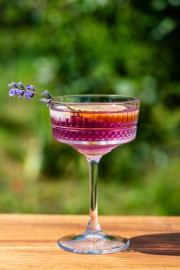 Yeşil bir yaz bahçesine karşı ahşap masada zarif kristal bardakta servis edilen parlak kokteyl ya da limonata. Eflatun çiçekli mor renkli içecek ve bir dilim limon.