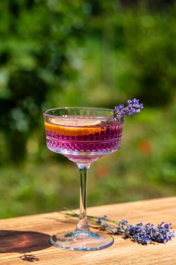 Yeşil bir yaz bahçesine karşı ahşap masada zarif kristal bardakta servis edilen parlak kokteyl ya da limonata. Eflatun çiçekli mor renkli içecek ve bir dilim limon.