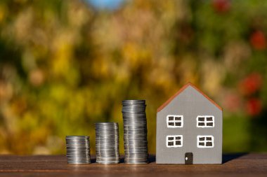 Küçük oyuncak ahşap ev modeli ve ahşap masa üzerinde bozuk para yığını, açık havada, kapat. Konut, ipotek ve gayrimenkul yatırımının satın alınması veya inşası için para biriktirme fikri