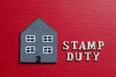 Inscription stamp duty made of wooden letters next gray wooden house on a red background, close up. Stamp Duty is a tax levied on the purchase of real estate or land, as well as on other transactions