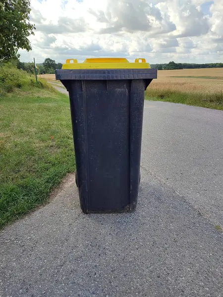 Dark garbage can with a yellow lid for plastic waste stands alone on the side of the road on a field path