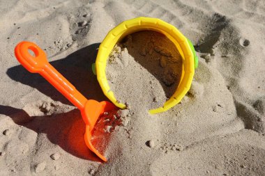 Kuma saplanmış küçük turuncu plastik bir kürek, kum havuzunda kum dolu sarı bir plastik kovanın yanında.