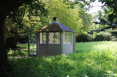 Camdan pencereleri olan küçük, güzel bir ahşap çardak ağaçlarla çevrili yeşil bir bahçede kapısı açık duruyor.