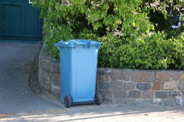Mavi çöp tenekesi, yeşil çitli tuğla bir duvara karşı duruyor.