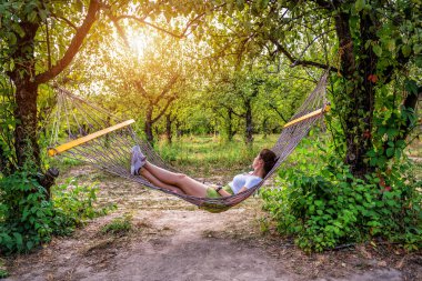 Young woman resting in comfortable hammock at green summer orchard garden