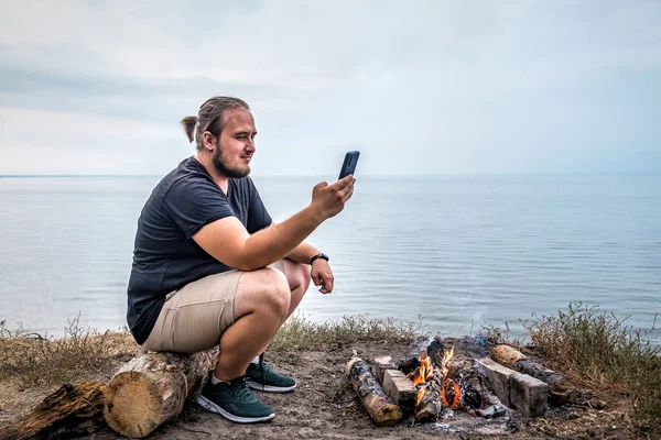 Jovem Adulto Sentado Margem Lago Perto Fogueira Com Seu Smartphone — Fotografia de Stock