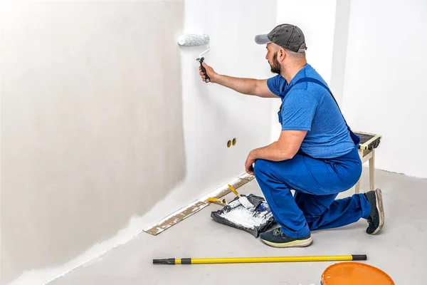 Builder in blue painting the wall with painting roller in white color. Industrial theme