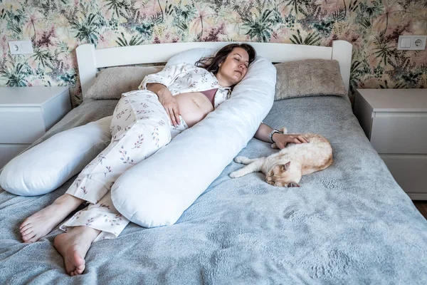 Pregnant woman sleeping in body pillow with red cat in bedroom