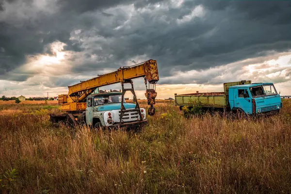 Abandoned construction mobile crane truck and old truck under stormy clouds at sunset