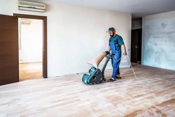 Professional carpenter worker in blue overalls grinding sanding wooden parquet floor by using floor sander. Industrial theme