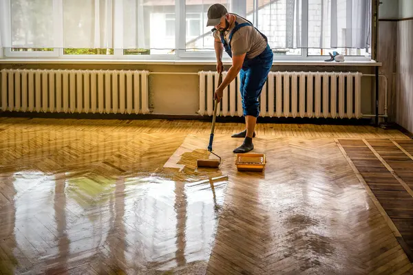 Professional worker in blue overalls lacquering varnishing old parquet floors using roller. Floor renovation