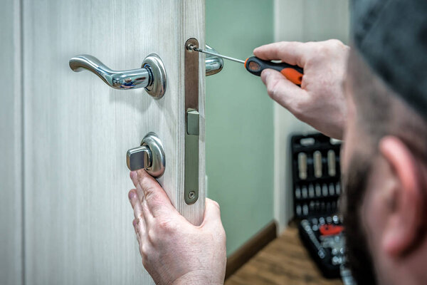 Professional locksmith worker installing the part of lock in wooden door by the screwdriver. Industrial theme
