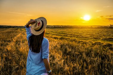 Buğday tarlasında gün batımında ya da gün doğumunda hasır şapkalı ve mavi gömlekli kadın.