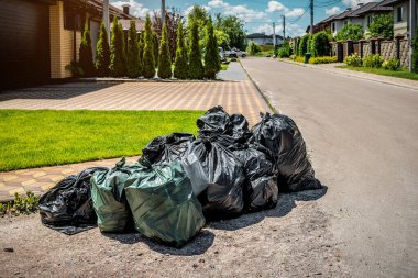 Asfalt bir sokakta, güneş ışığı altında yeşil çimlerin yanında siyah plastik çöp torbaları.
