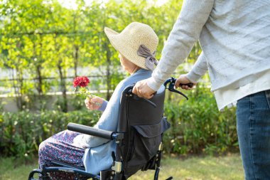Bakıcı kız, parkta tekerlekli sandalyede kırmızı gül tutan Asyalı yaşlı ya da yaşlı kadına sarılıp yardım ediyor..