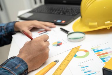 Asian engineer and partner team learn and talk with house plan project blueprint, graph and yellow helmet and engineering construction tools on table work.