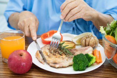 Asyalı yaşlı ya da yaşlı kadın hasta somon bifteği ve sebzeli sağlıklı yemek yerken hastanede aç ve aç bir şekilde oturuyor..