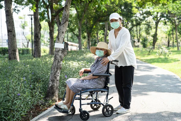 Caregiver help and care Asian senior or elderly old lady woman patient sitting on wheelchair and wear a face mask to travel in park,