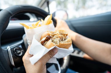 Arabada yemek için hamburger ve patates kızartması tutan Asyalı kadın tehlikeli ve kaza riski taşıyor..