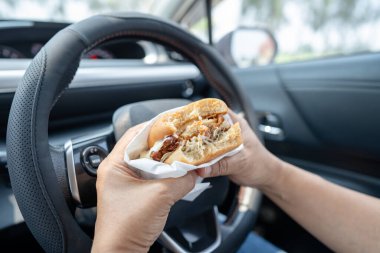 Arabada yemek için hamburger tutan Asyalı kadın tehlikeli ve kaza riski taşıyor..