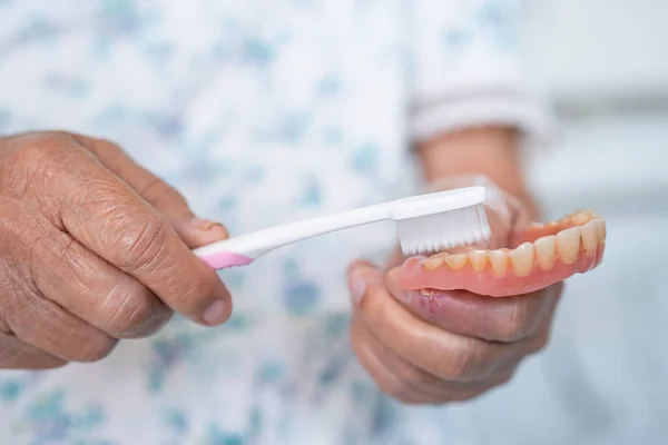 Asian elderly woman patient use toothbrush to clean partial denture of replacement teeth.