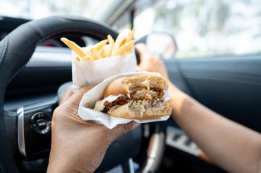 Arabada yemek için hamburger ve patates kızartması tutan Asyalı kadın tehlikeli ve kaza riski taşıyor..