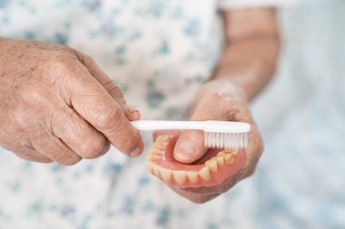 Asyalı yaşlı kadın hasta diş fırçasıyla takma diş protezini temizliyor..