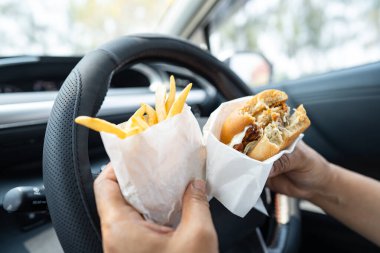 Arabada yemek için hamburger ve patates kızartması tutan Asyalı kadın tehlikeli ve kaza riski taşıyor..