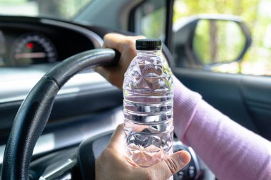 Asyalı kadın sürücü araba sürerken su içmek için şişe tutuyor. Plastik sıcak su şişesi ateşe neden olur..