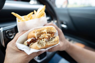 Arabada yemek için hamburger ve patates kızartması tutan Asyalı kadın tehlikeli ve kaza riski taşıyor..