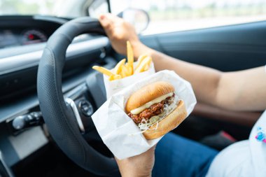 Arabada yemek için hamburger ve patates kızartması tutan Asyalı kadın tehlikeli ve kaza riski taşıyor..