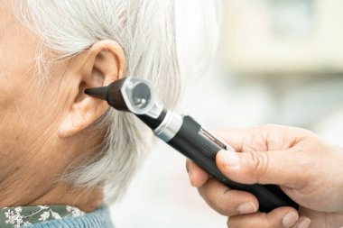 Odiyolog veya ENT doktoru otoskop kullanarak işitme sorunu olan Asyalı yaşlı kadın hastaların kulaklarını kontrol eder..