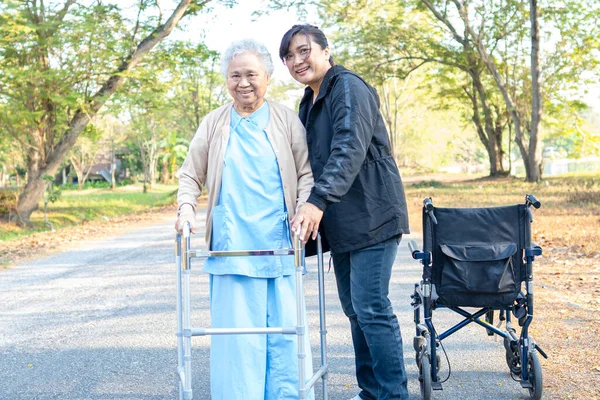 Caregiver help Asian senior woman walk with walker with wheelchair at park.