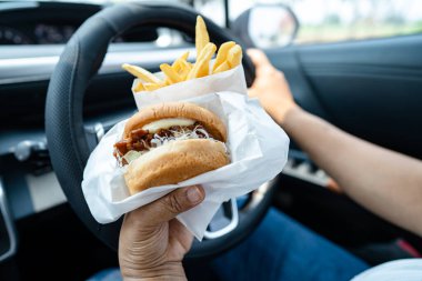 Arabada yemek için hamburger ve patates kızartması tutan Asyalı kadın tehlikeli ve kaza riski taşıyor..