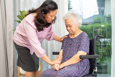 Bakıcı, tekerlekli sandalyedeki Asyalı yaşlı kadına evde sevgiyle yardım ediyor..