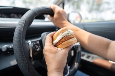 Arabada yemek için hamburger tutan Asyalı kadın tehlikeli ve kaza riski taşıyor..