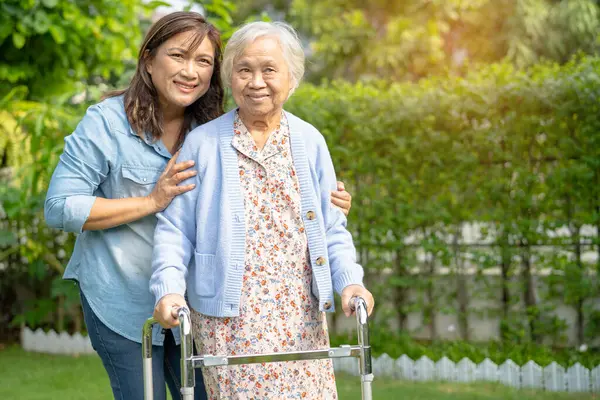 Doctor caregiver help and care Asian senior woman patient walk with walker in park at hospital.