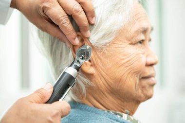 Odiyolog veya ENT doktoru otoskop kullanarak işitme sorunu olan Asyalı yaşlı kadın hastaların kulaklarını kontrol eder..
