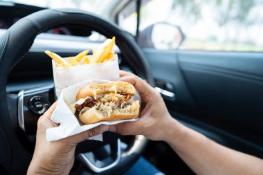 Arabada yemek için hamburger ve patates kızartması tutan Asyalı kadın tehlikeli ve kaza riski taşıyor..