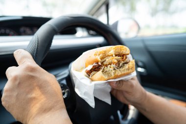 Arabada yemek için hamburger ve patates kızartması tutan Asyalı kadın tehlikeli ve kaza riski taşıyor..